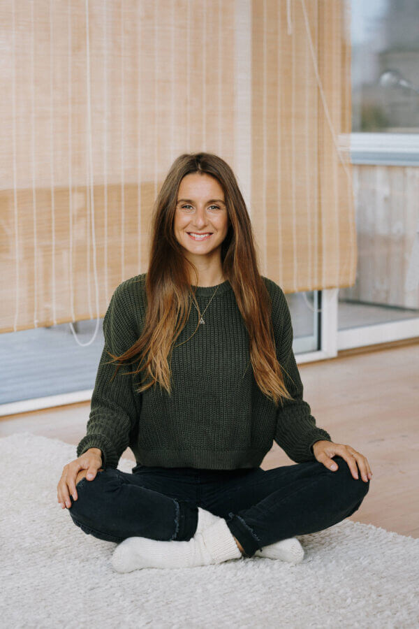 Testimonial – Laura Malina Seiler (Abschlussfeier) Laura Malina Seiler sitzt lächelnd im Schneidersitz auf einem Teppich in einem hellen Raum.