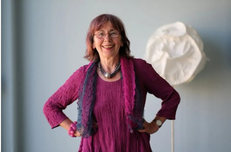 Testimonial – Margret Rasfeld (Fundraising Event) Margret Rasfeld steht lächelnd in einem violetten Kleid vor hellem Hintergrund